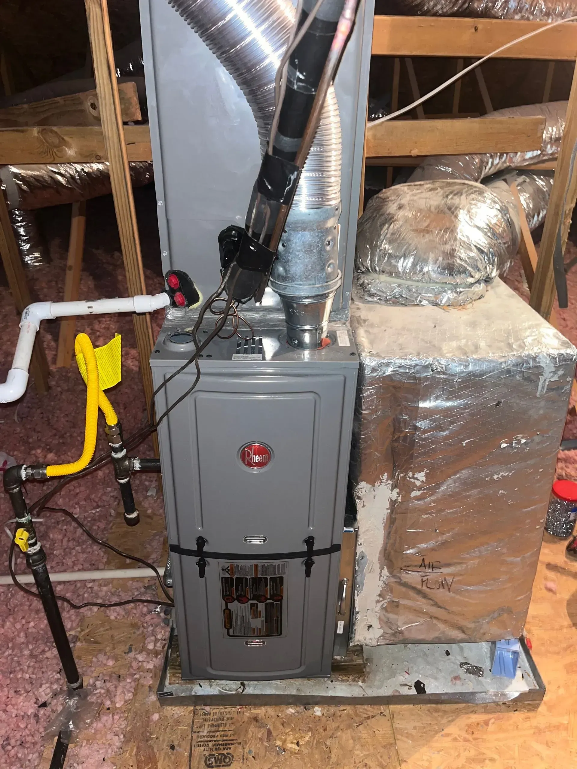 Gray Rheem furnace in an attic. Ductwork and gas line visible.
