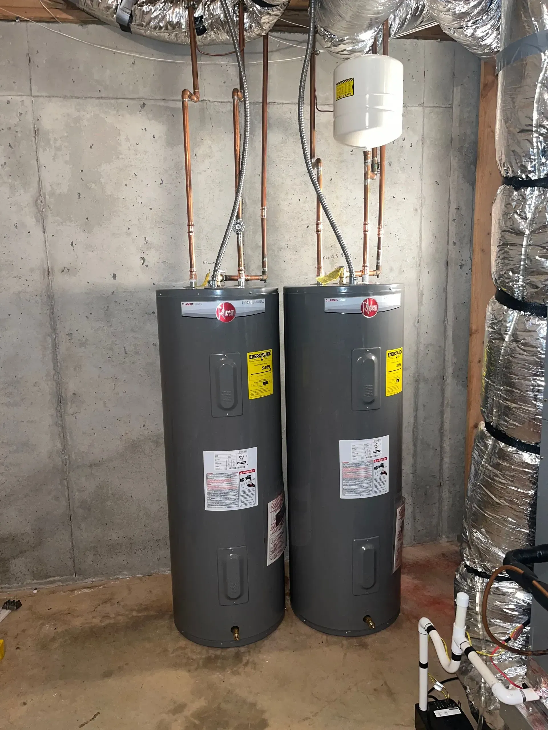 Two tall gray water heaters in a concrete basement, copper pipes, expansion tank.