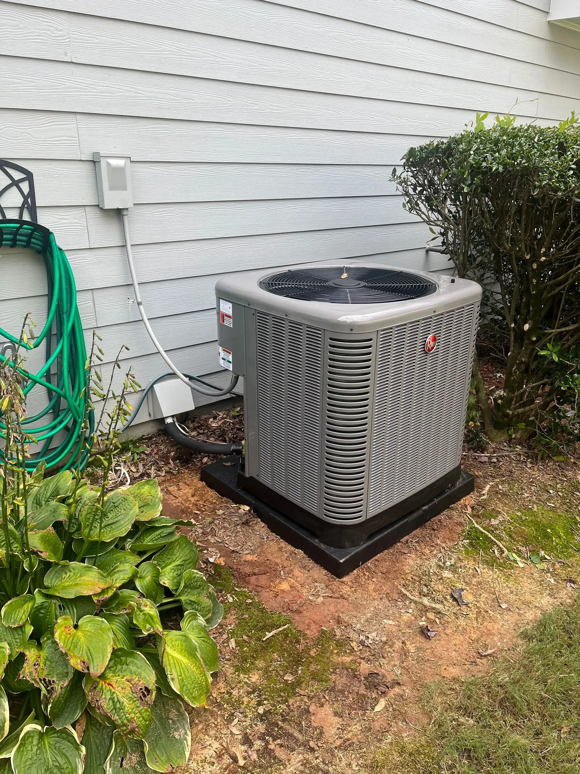 Gray AC unit sits on a black base near a house and greenery, with a hose nearby.