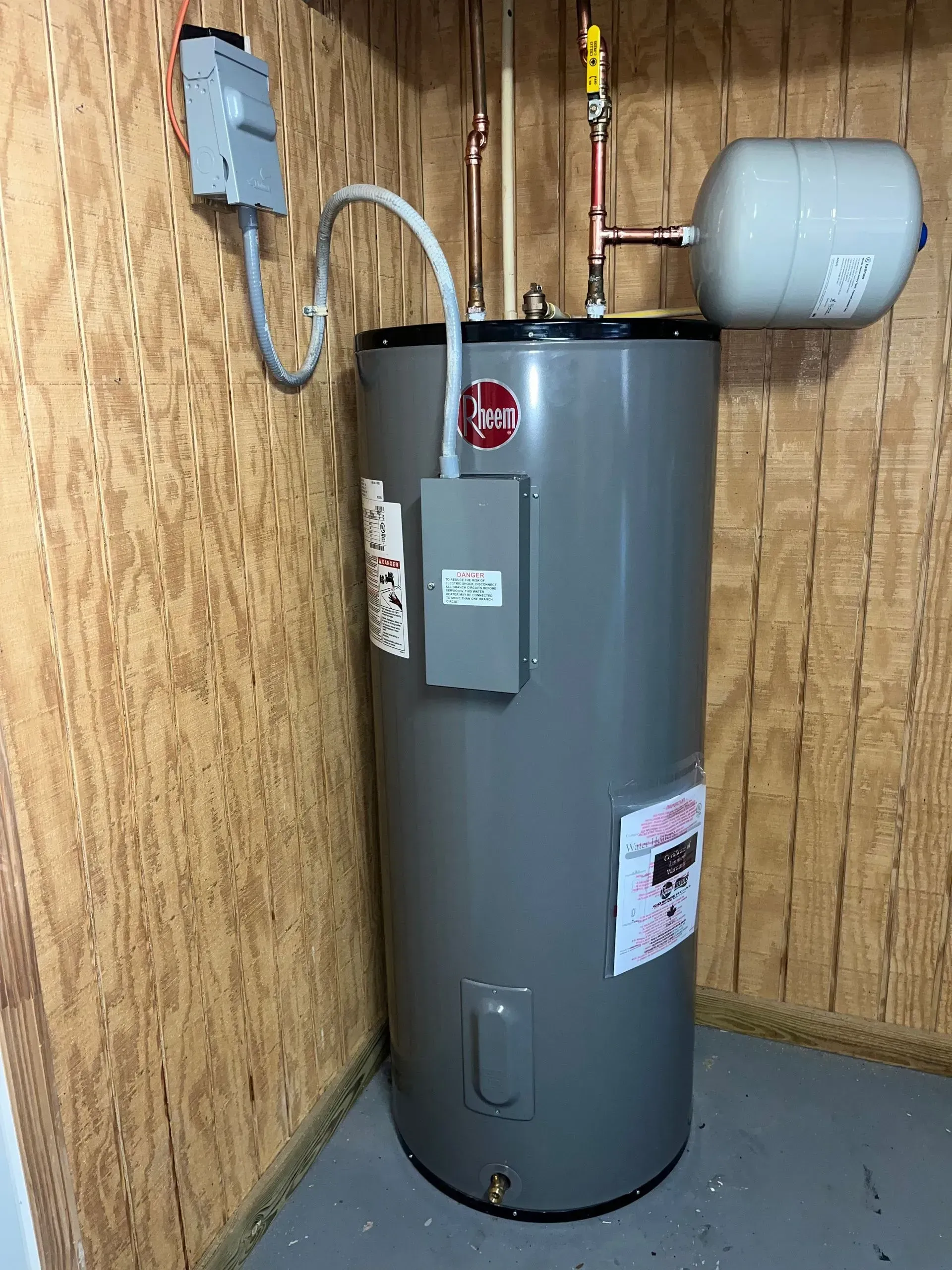 A tall, gray water heater in a corner of a room with wood-paneled walls, with plumbing and electrical connections visible.