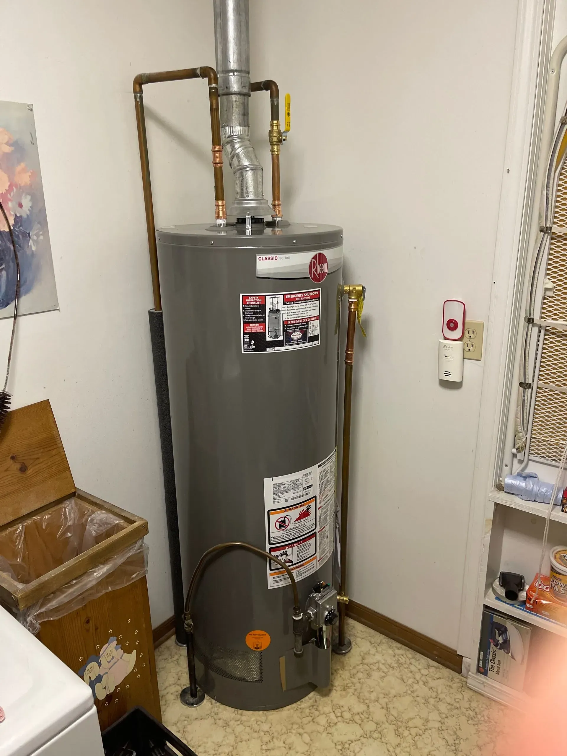 A tall gray water heater with copper pipes and a silver vent pipe sits in a corner near a washer and a wooden box.