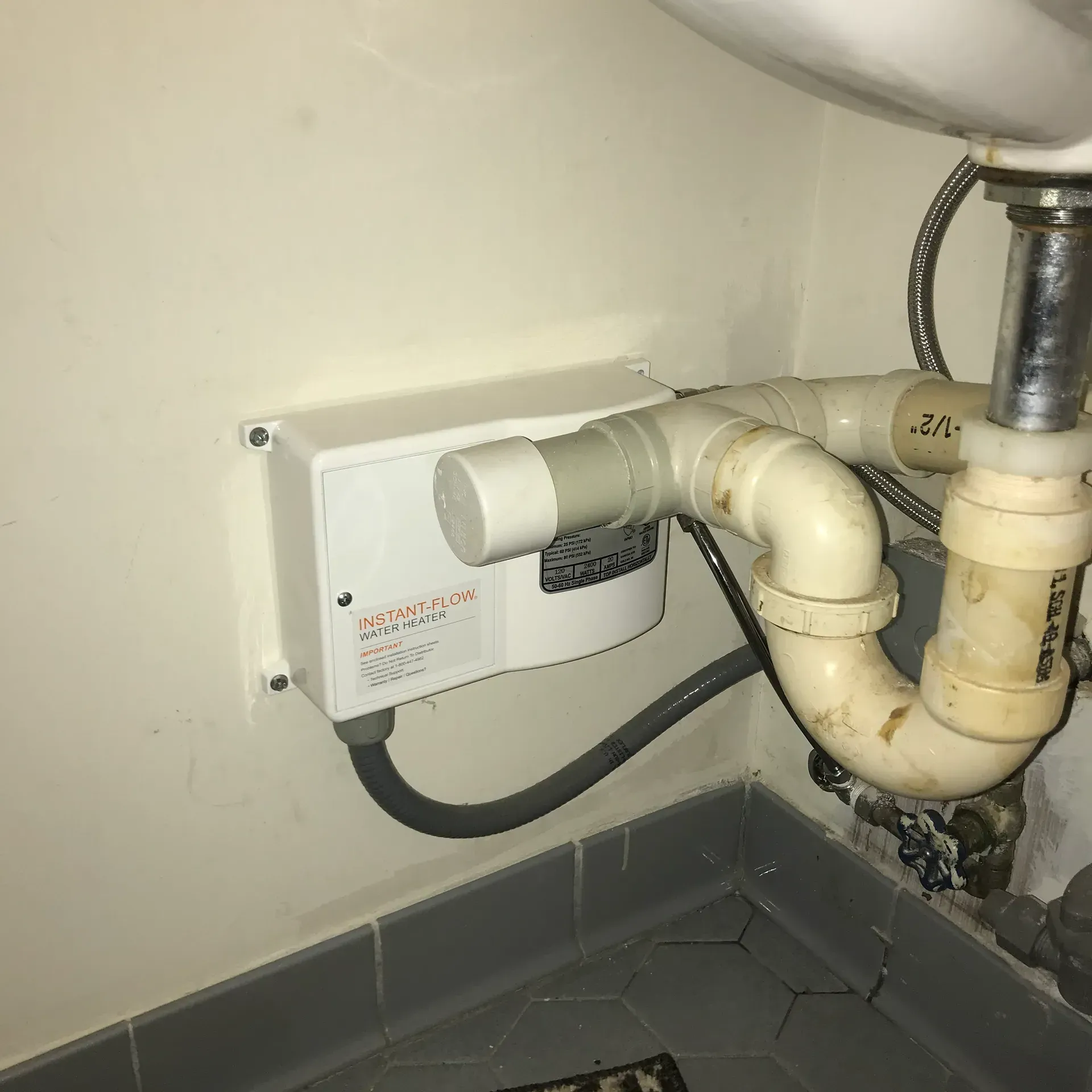 White appliance mounted on a wall next to sink plumbing. Gray pipe, tan wall and black floor.