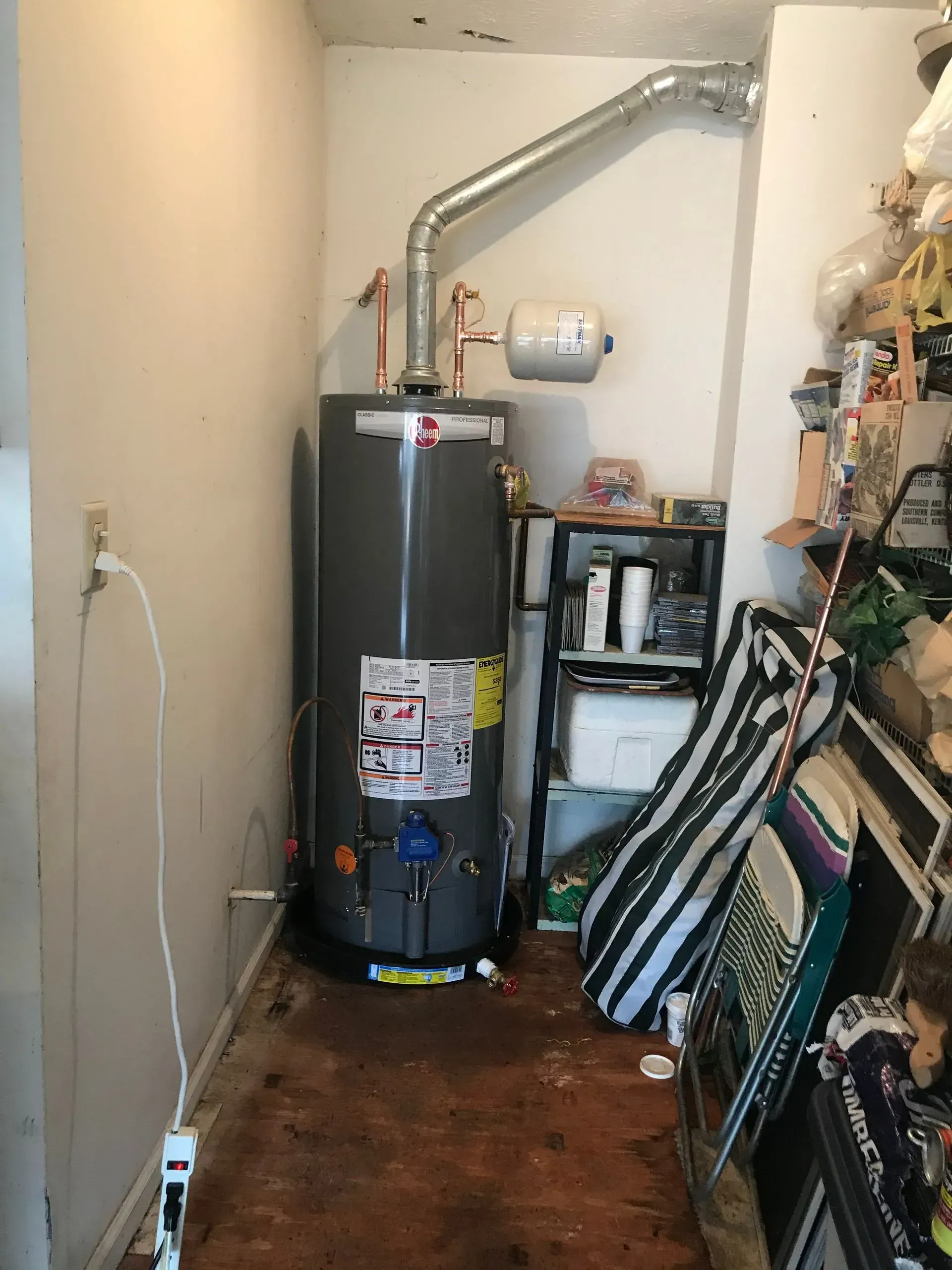 Water heater in a narrow room with vent, pipes, and cluttered shelving.