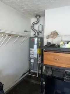 Water heater in a garage, with silver vent pipe, next to a wooden cabinet and shelving.