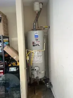 A tall, silver water heater is installed in a narrow, white-walled space in a garage.