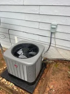 Gray Rheem air conditioner unit near a light gray house. A black conduit goes to an electrical outlet.