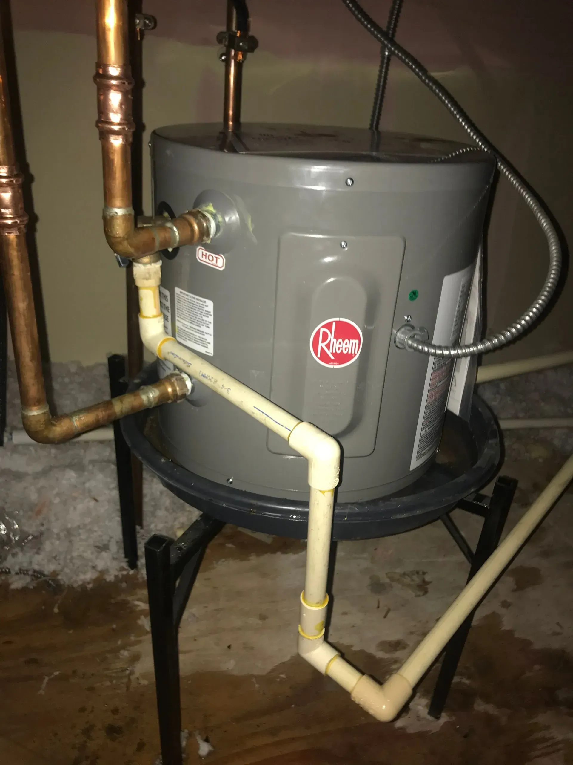 Gray Rheem water heater on a black stand, with copper and PVC pipes, and water on the floor.