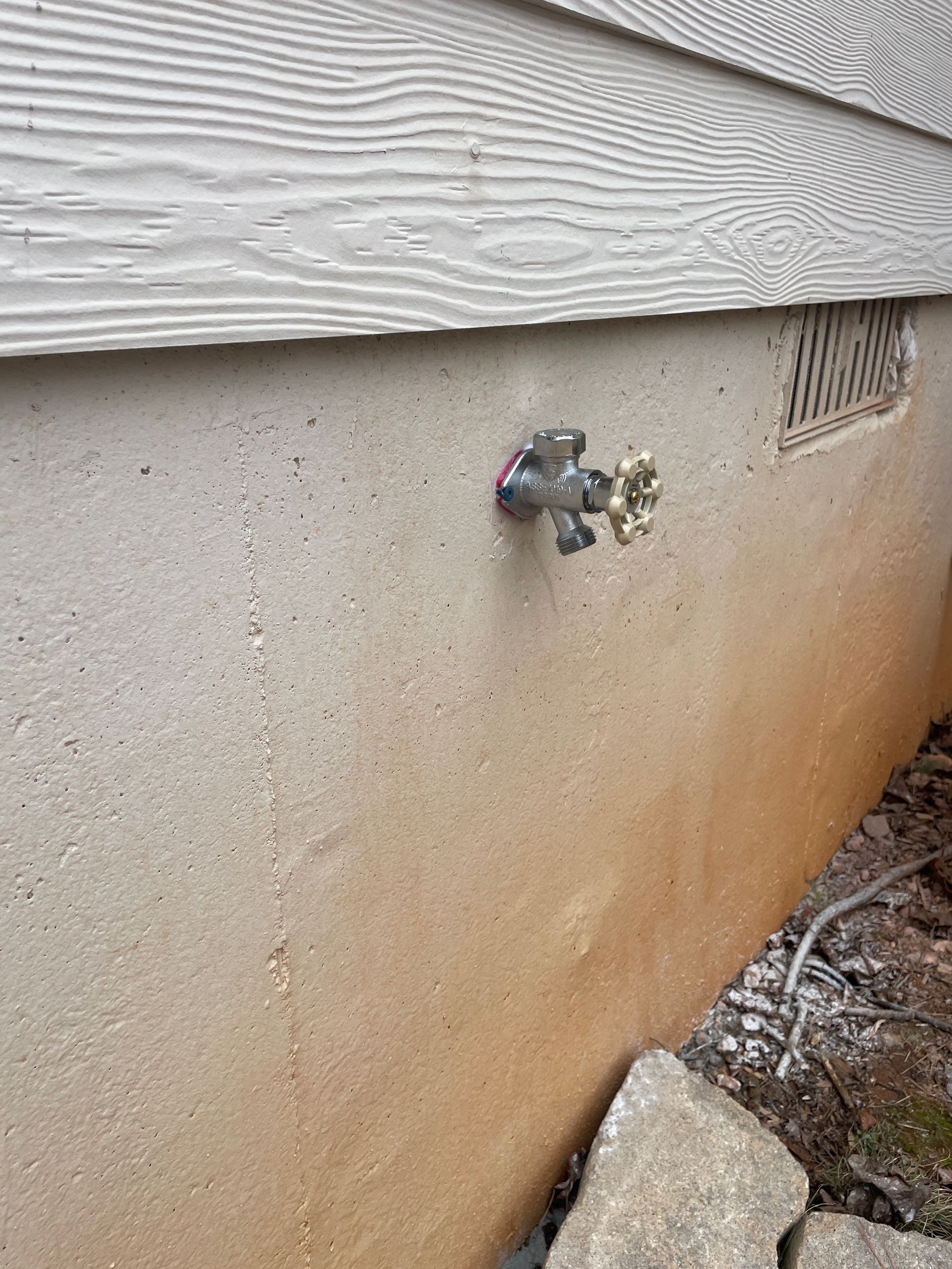 Outdoor spigot on a light brown stucco foundation below white siding.