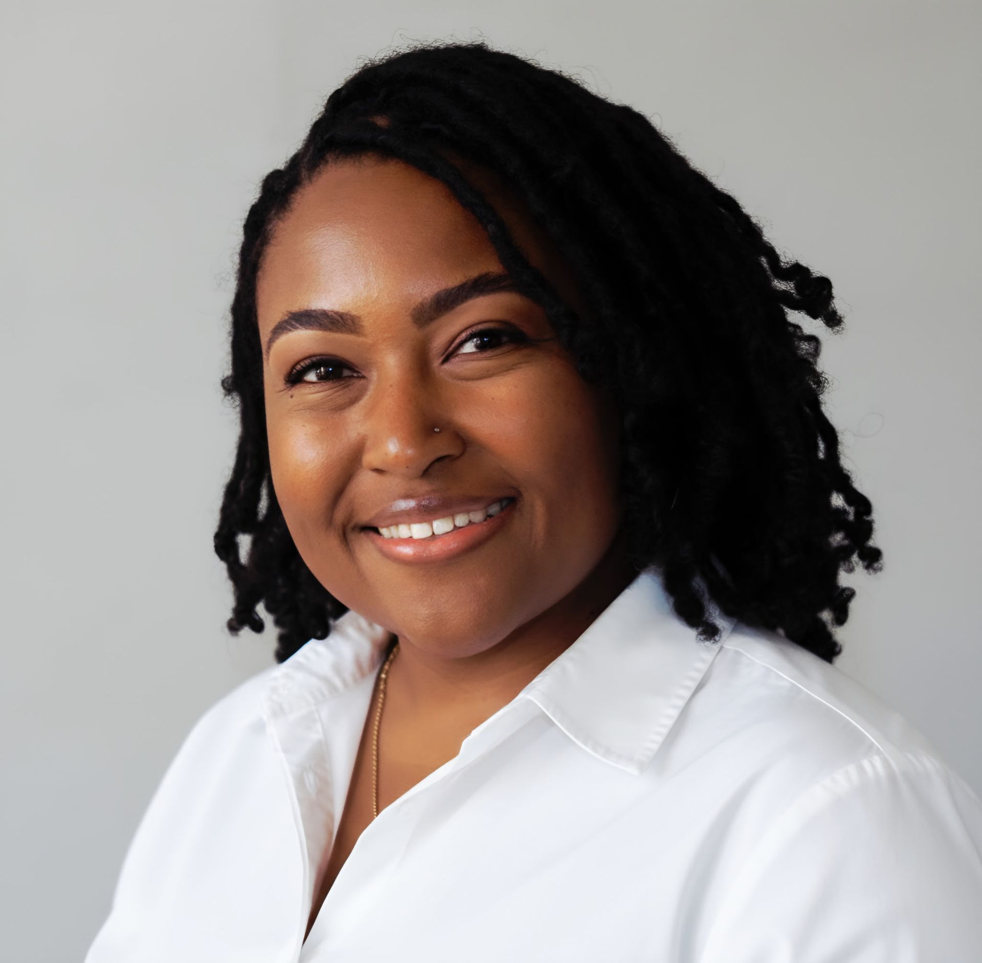 Smiling person with shoulder-length black hair in a white shirt against a light gray background