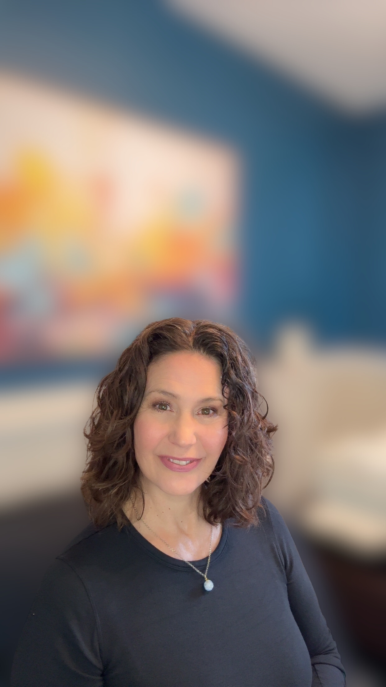 Woman with curly brown hair, smiling, wearing black top, background with art, blue wall.