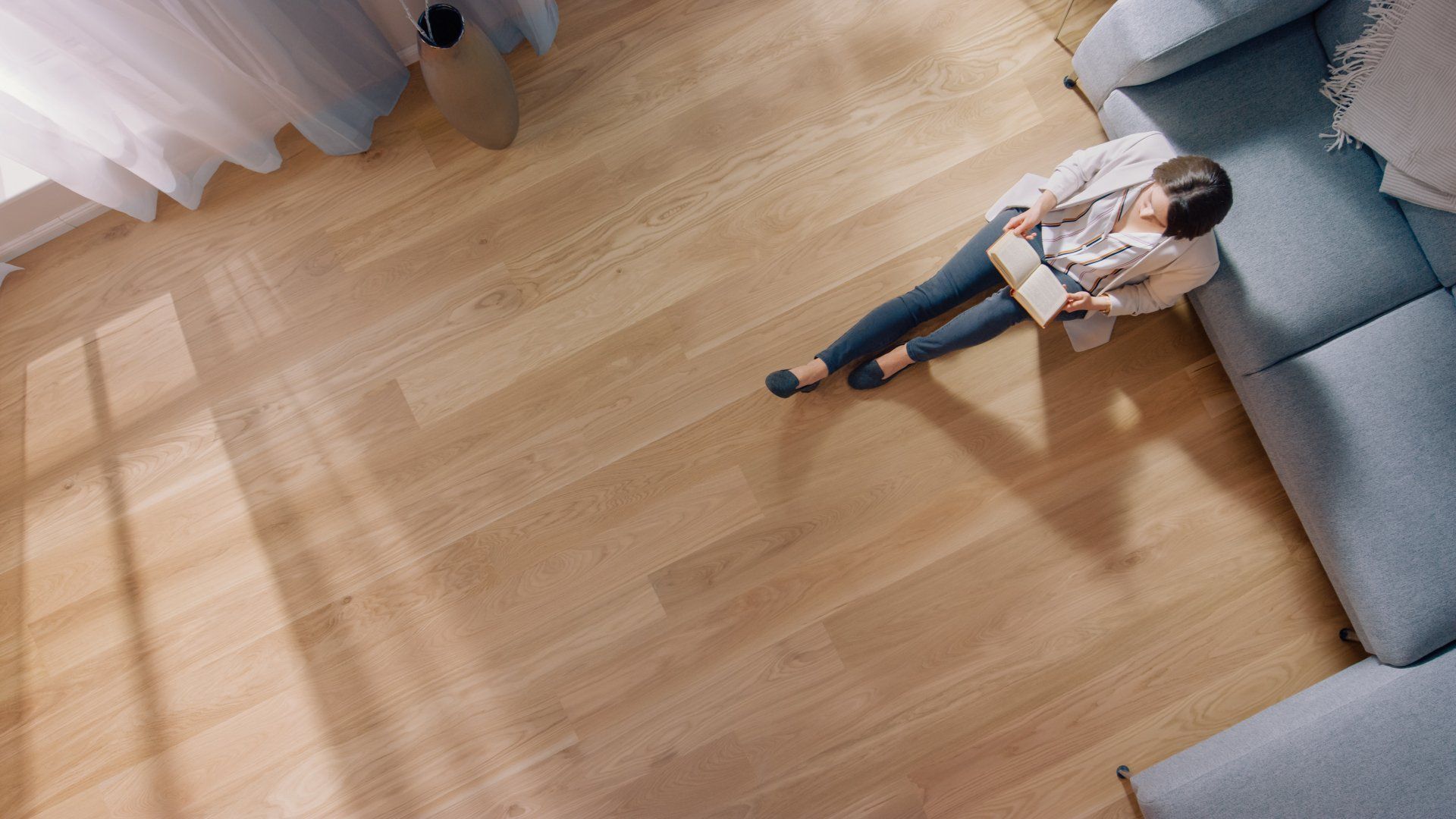 Young Woman is Sitting on a Floor and Reading a Book. Cozy Living Room with Modern Interior, Grey Sofa and Wooden Flooring. Top View Camera Shot.