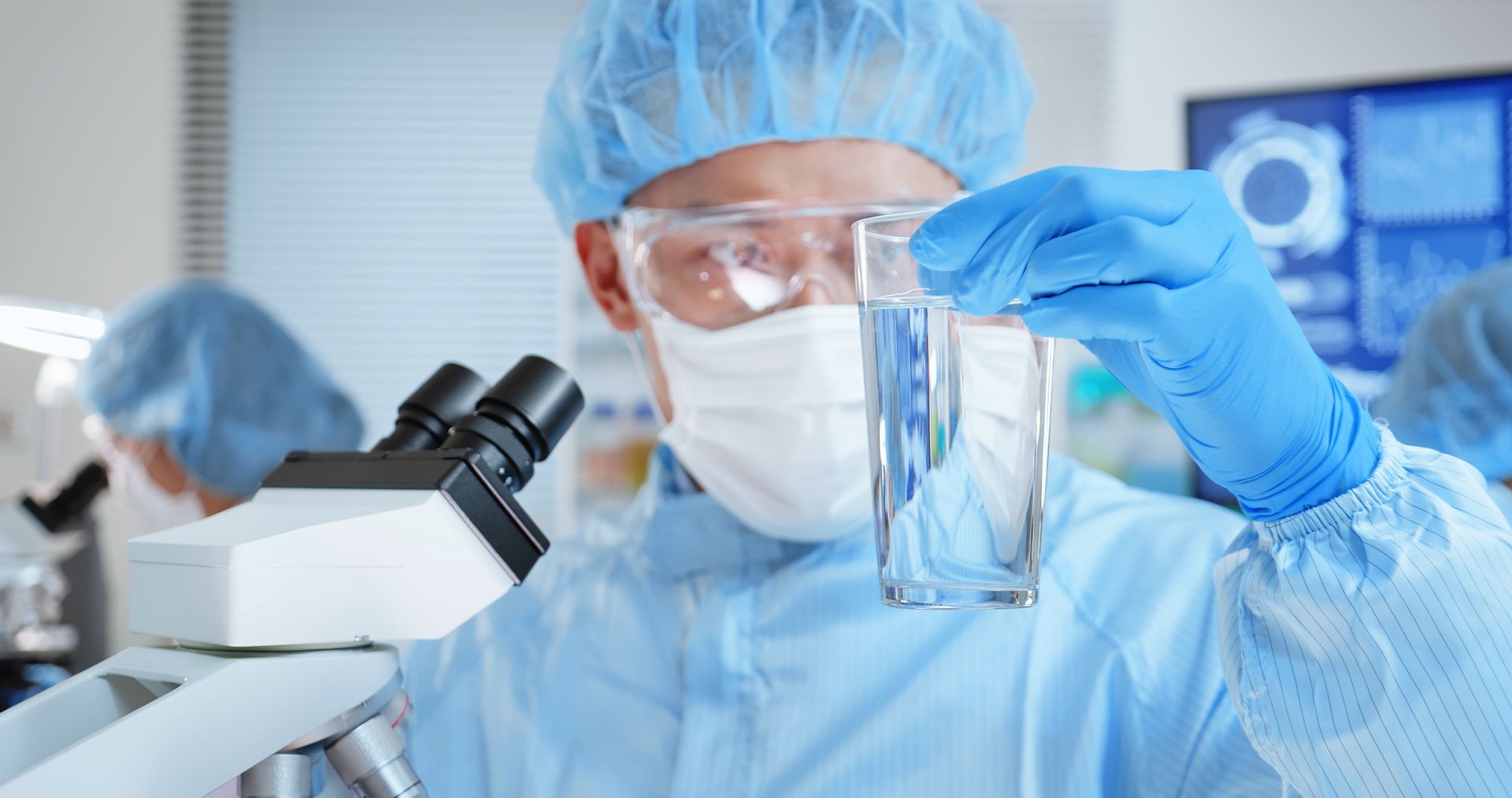 A scientist in PPE holds a water sample beaker in the lab. A scientist in PPE holds a water sample beaker in the lab.
