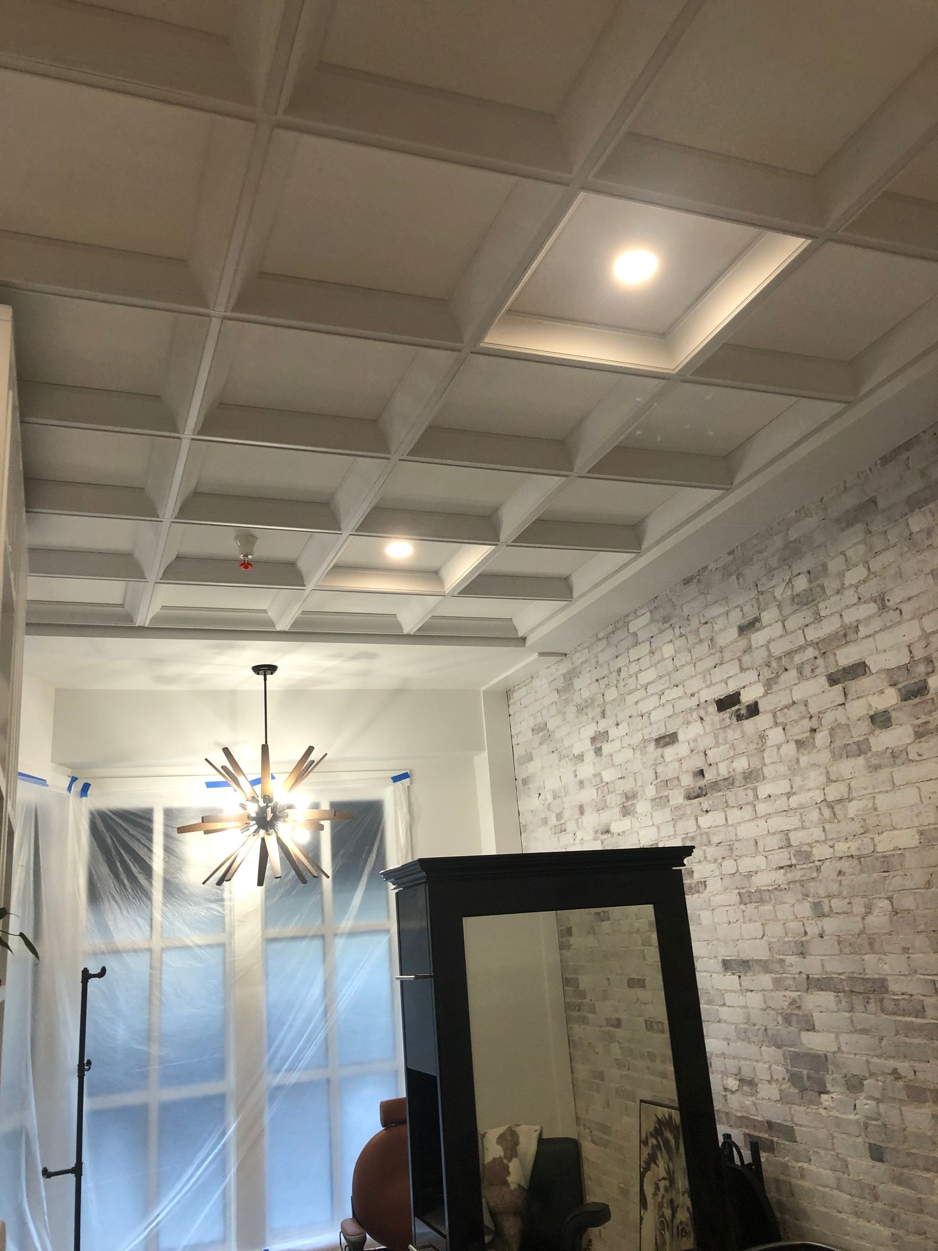 White coffered ceiling with recessed lights, above a room with exposed brick wall and a large window.