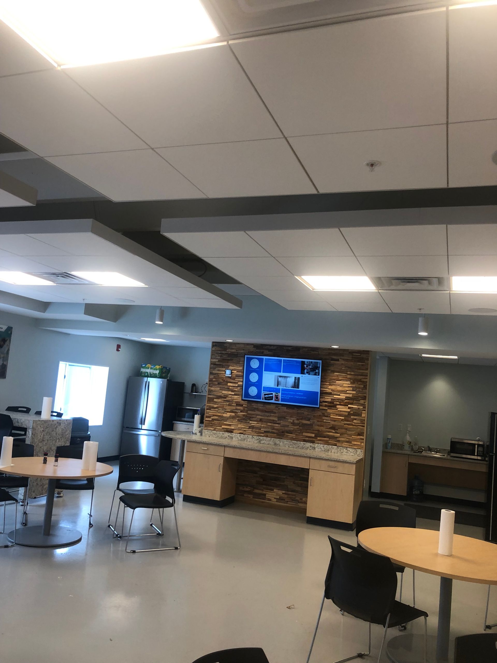 Indoor waiting area with tables, chairs, TV, and vending machine. Brick-patterned wall with built-in cabinet.