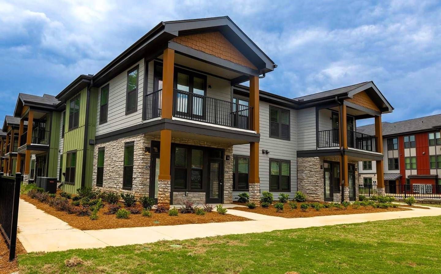 Modern apartment buildings with green, gray, and red facades, stone accents, and balconies under a cloudy sky.