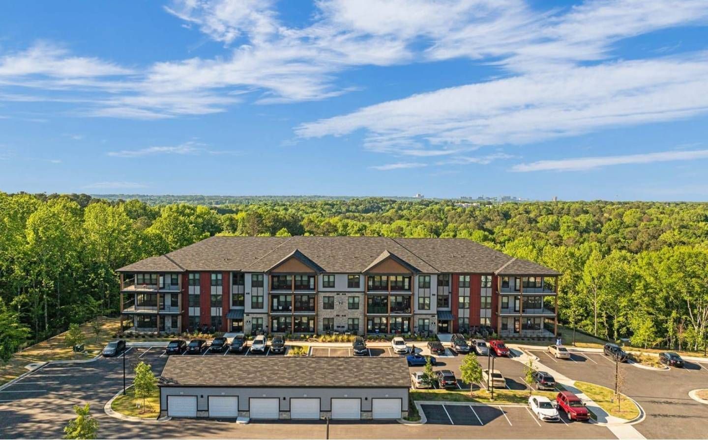 Apartment building with dark roof, red and gray accents, surrounded by trees; parking lot in front.