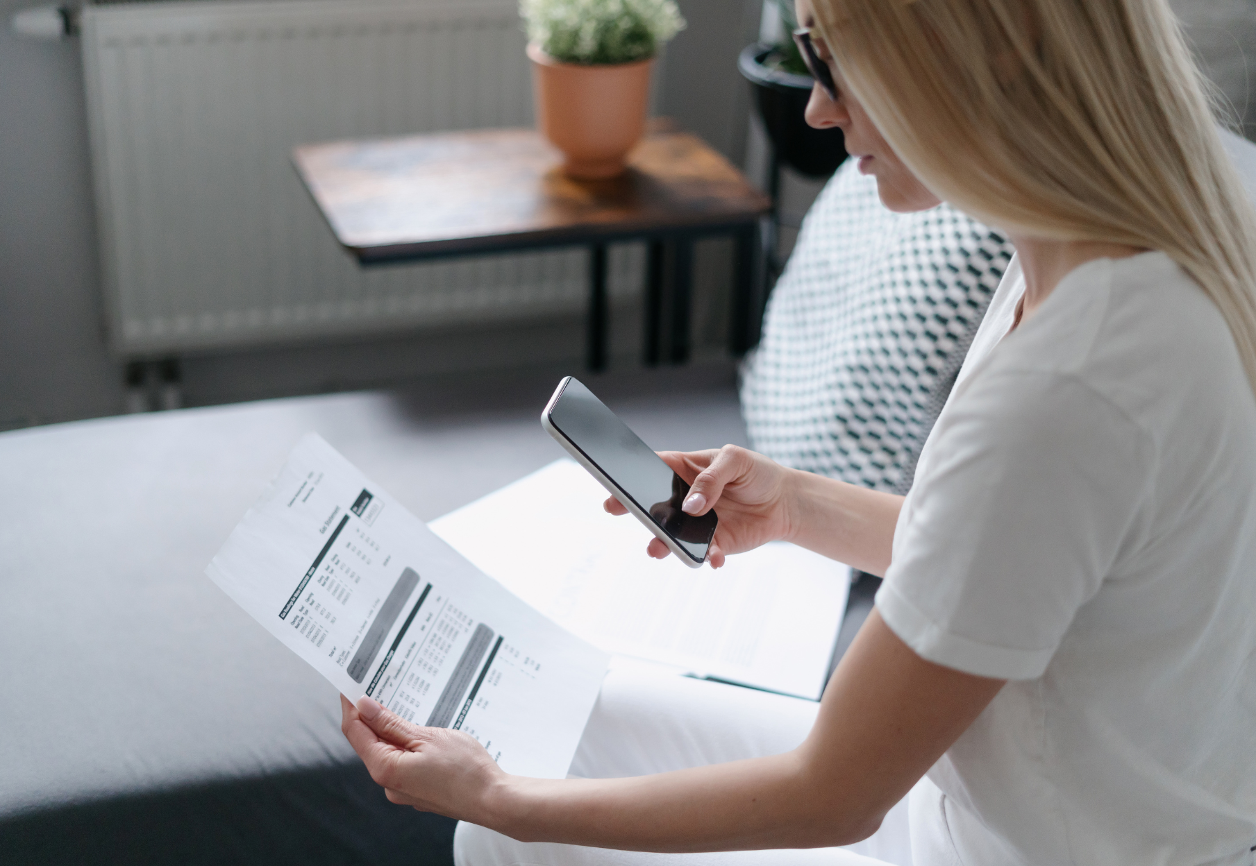 A woman is sitting on a couch holding a piece of paper and a cell phone.