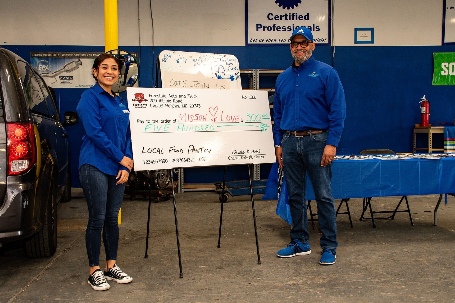 Two people hold a large check in a garage. They are smiling.