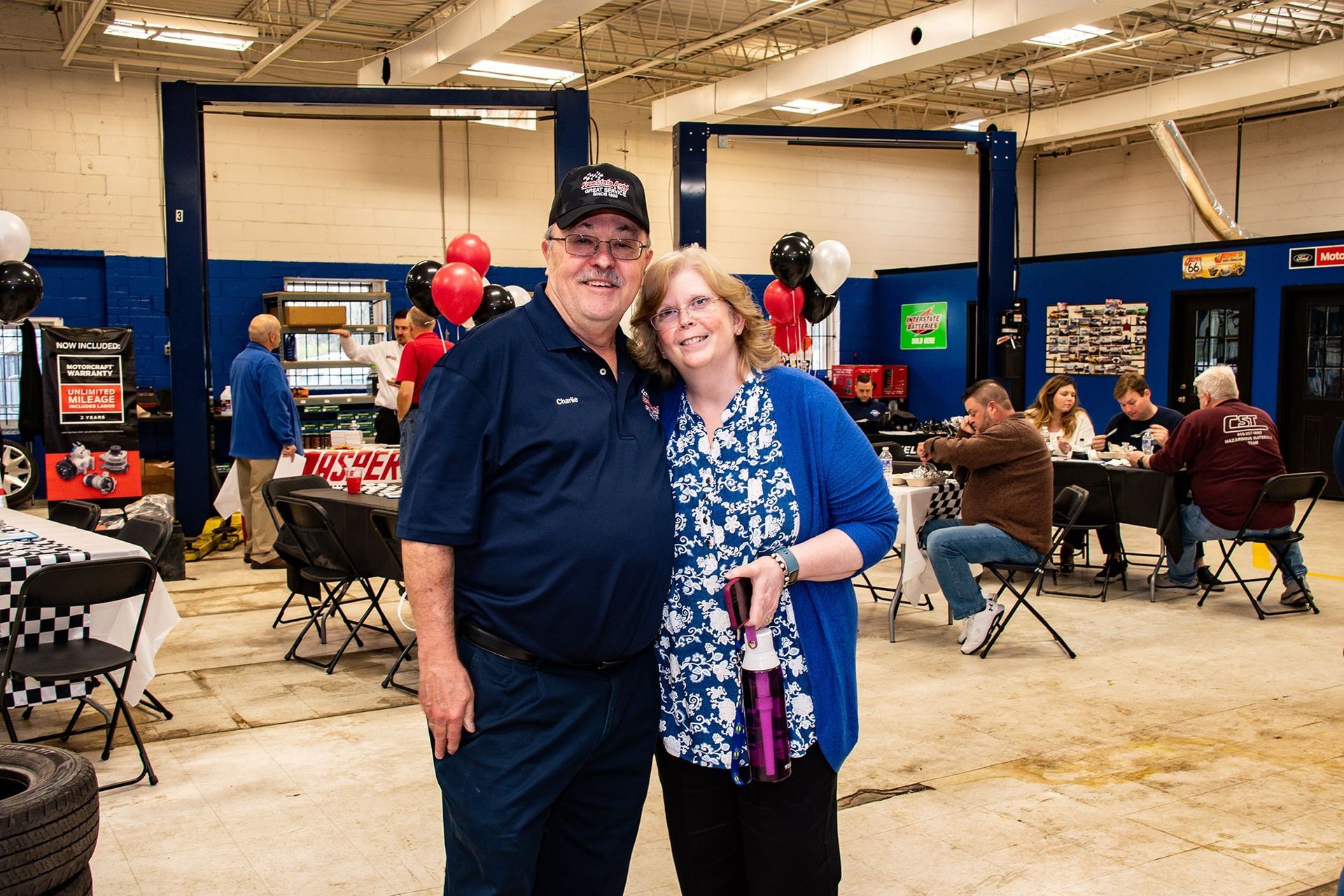 A couple smiles at a party in a garage with balloons, tables, and car lifts.