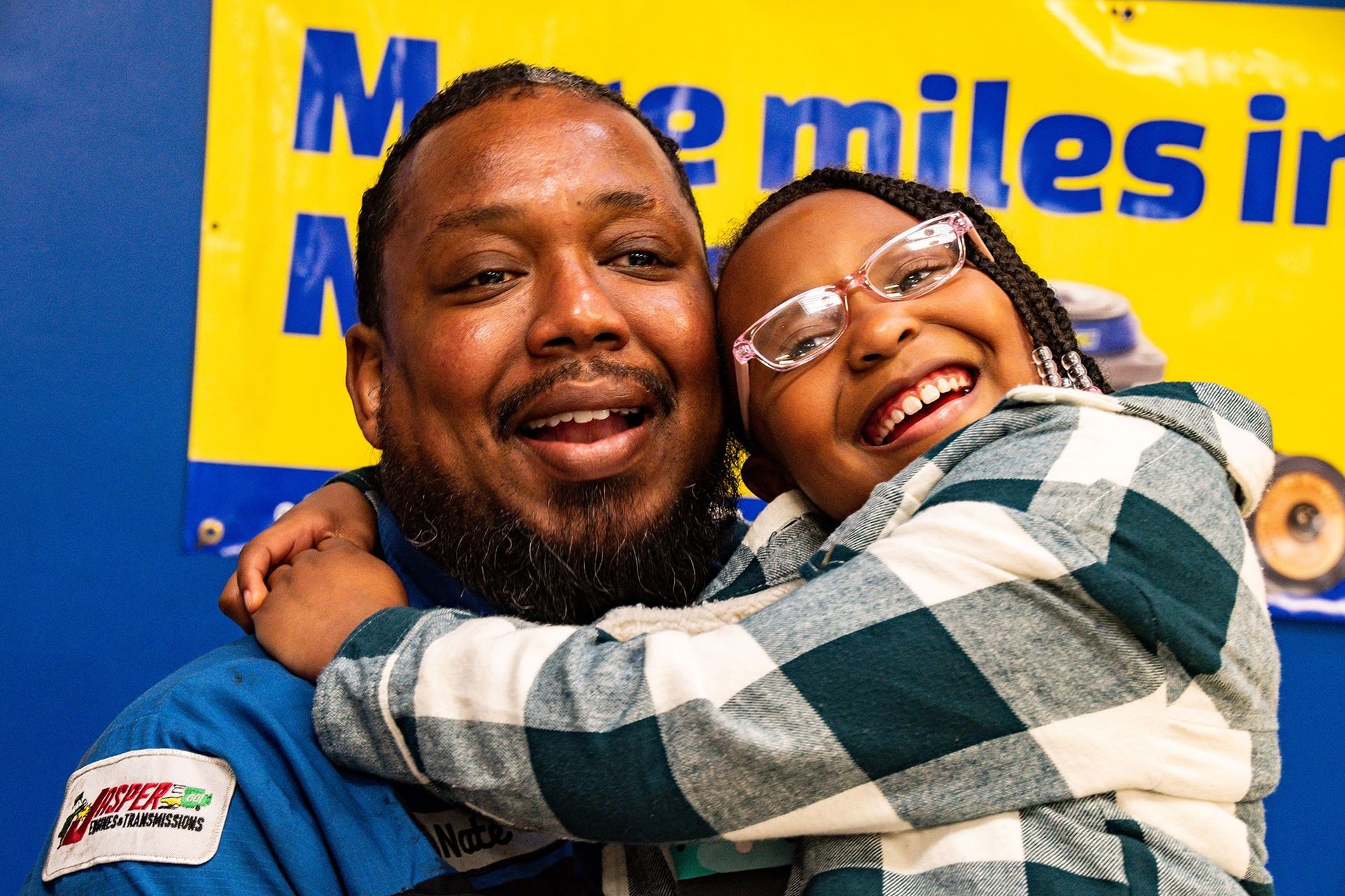 Man with a beard hugs a girl with glasses, both smiling, in front of a blue wall with a sign.