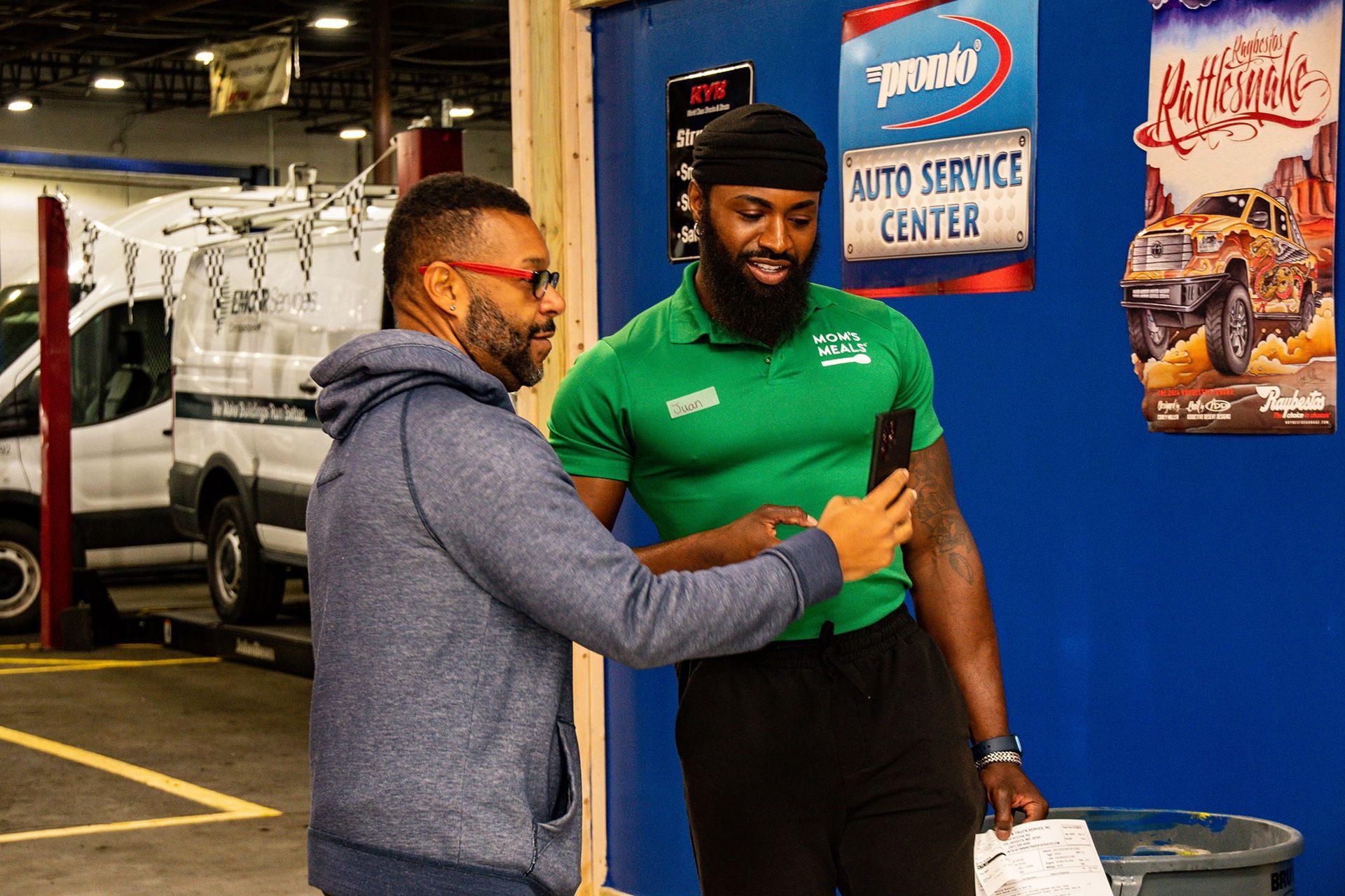 Two men in auto shop; one takes photo with phone, other smiles. Blue and green clothing.