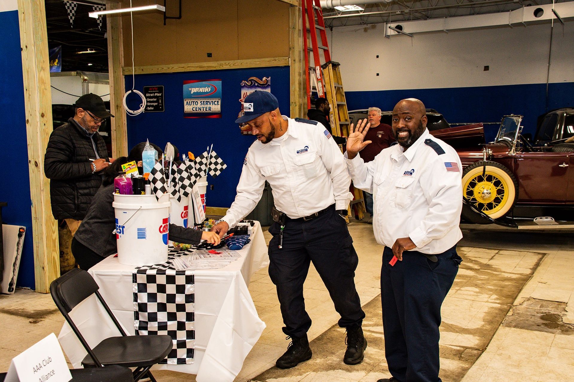Two men in work uniforms at a table with items, one waving, in a garage-like space.