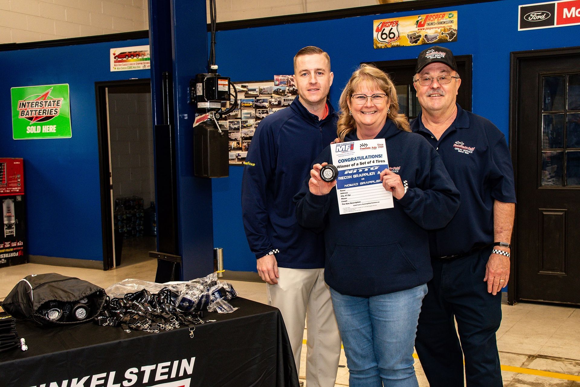 Three people in a garage holding an award and a part, smiling. A table displays more parts.