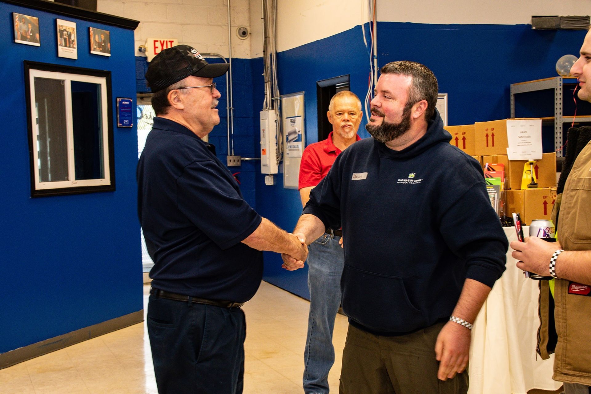Two men shaking hands in a blue-walled room. A third man in red shirt watches, a fourth to the right.
