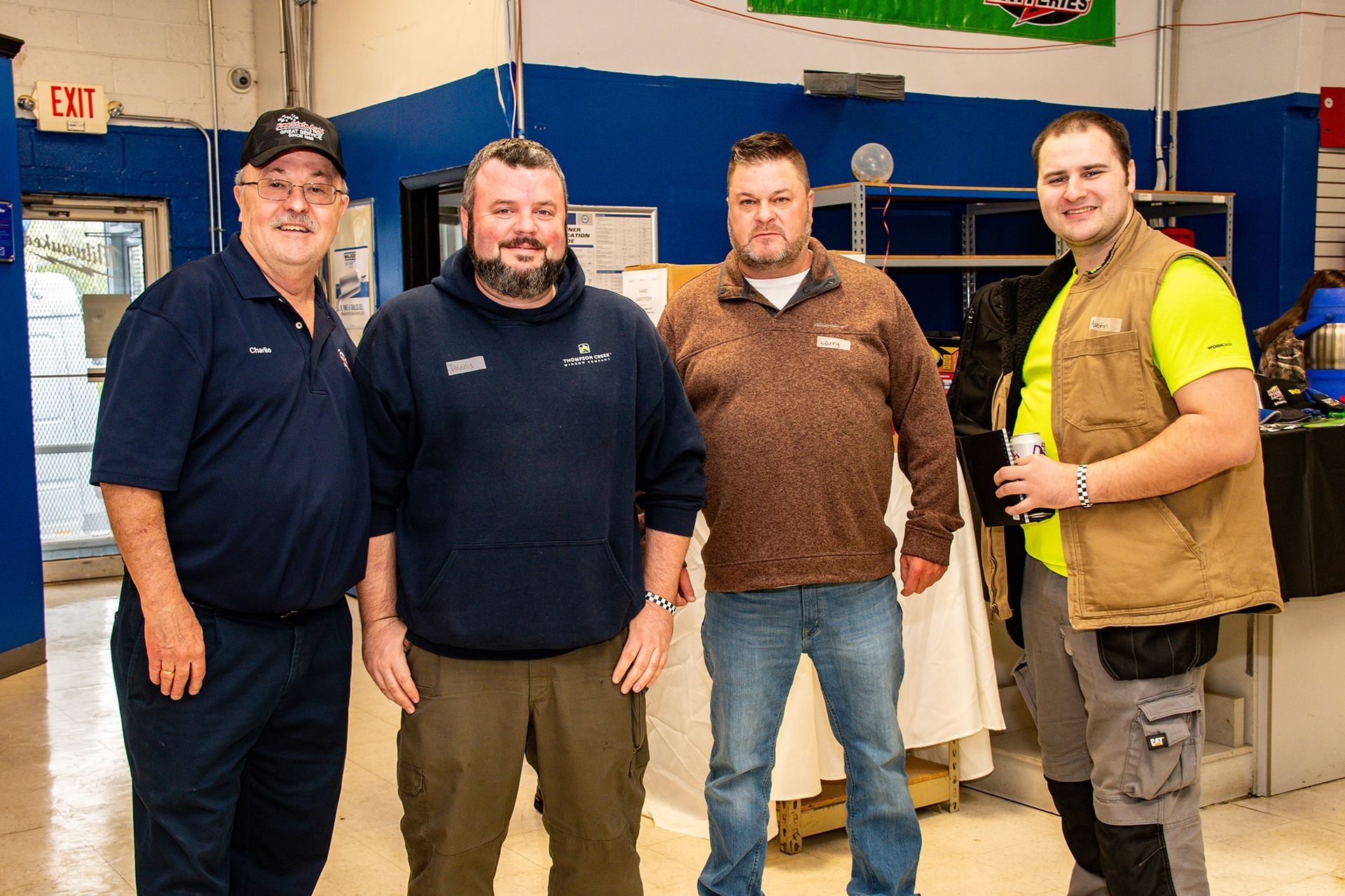Four men standing in a shop, smiling. One in brown sweater, another in yellow shirt with a vest. Blue and white walls in background.