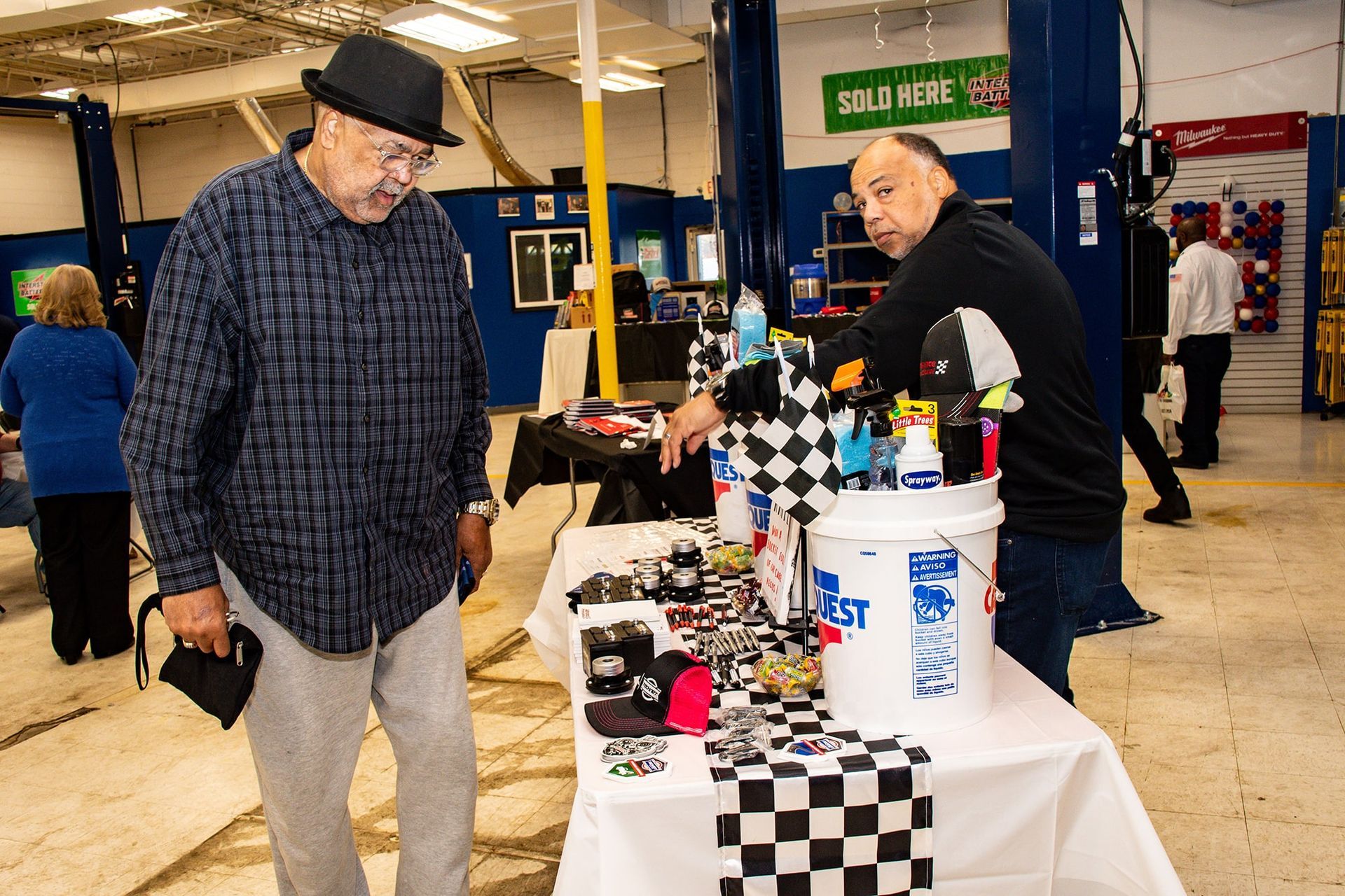 Two men at a checkered-table selling items in a garage-like setting. One man wears a fedora, the other points.