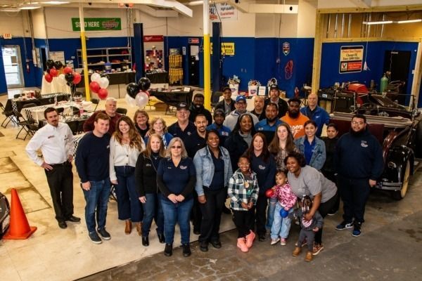 Group of people in an auto repair shop with a vintage car, balloons, and tables.
