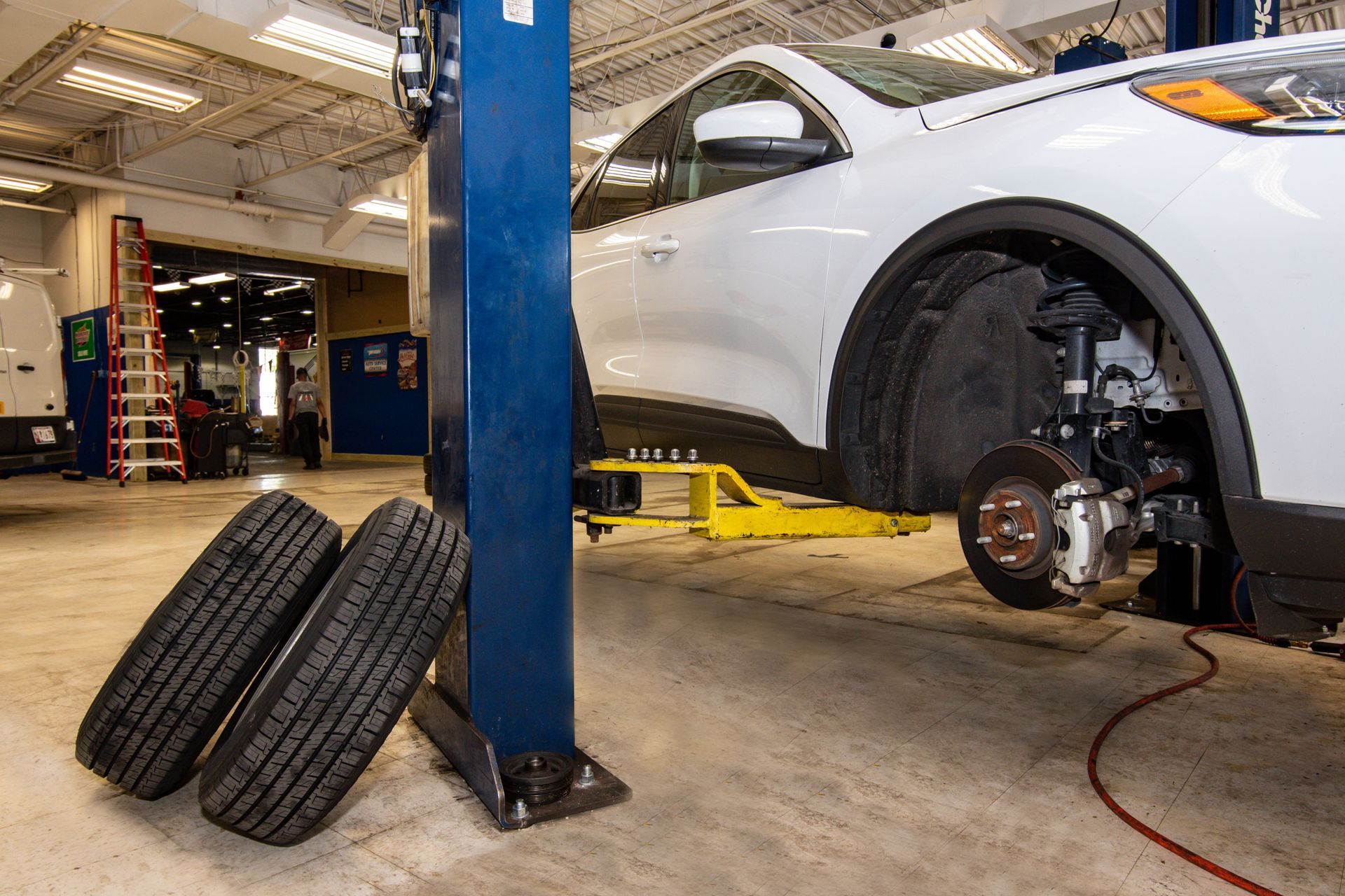 White car on a lift in a garage, tires leaning nearby. Brake work visible | Freestate Auto & Truck Service