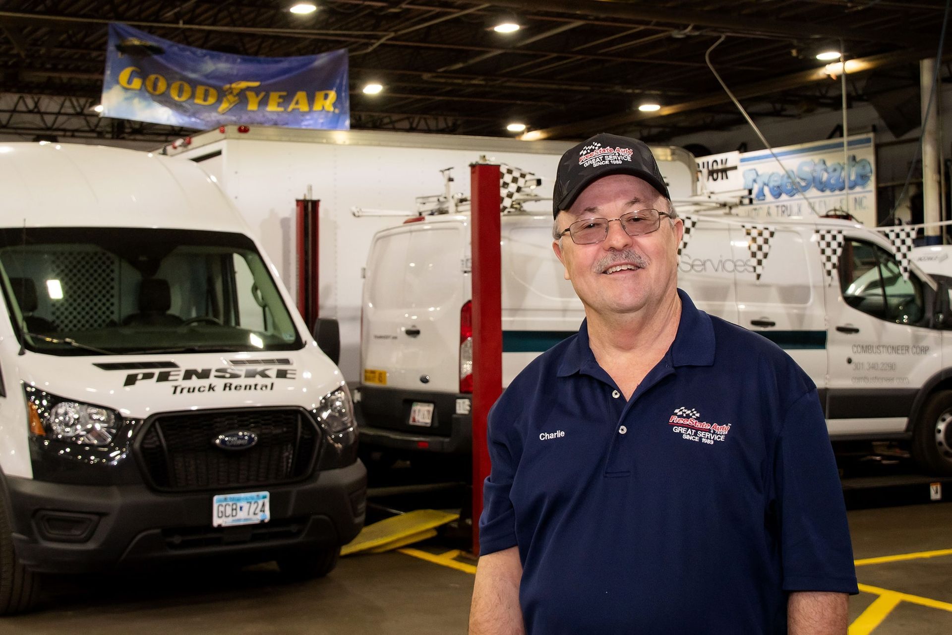 Man in a shop stands in front of vans; he wears a hat and shirt with a logo, Goodyear banner above.