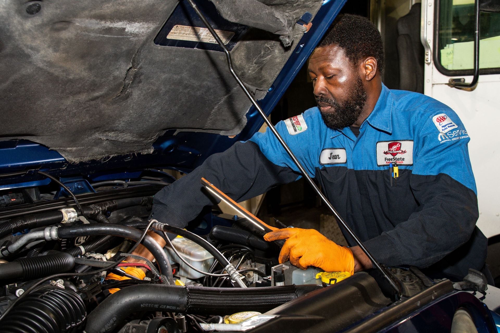 Mechanic in blue uniform inspects engine with a light. Orange gloves, dark skin, indoors | Freestate Auto & Truck Service
