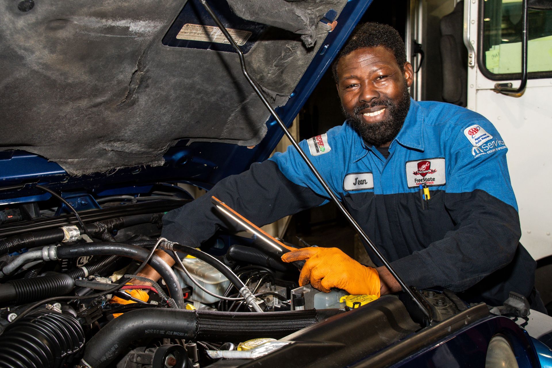 Mechanic working on an engine, smiling. Wearing gloves and blue work shirt, in an open engine bay | Freestate Auto & Truck Service