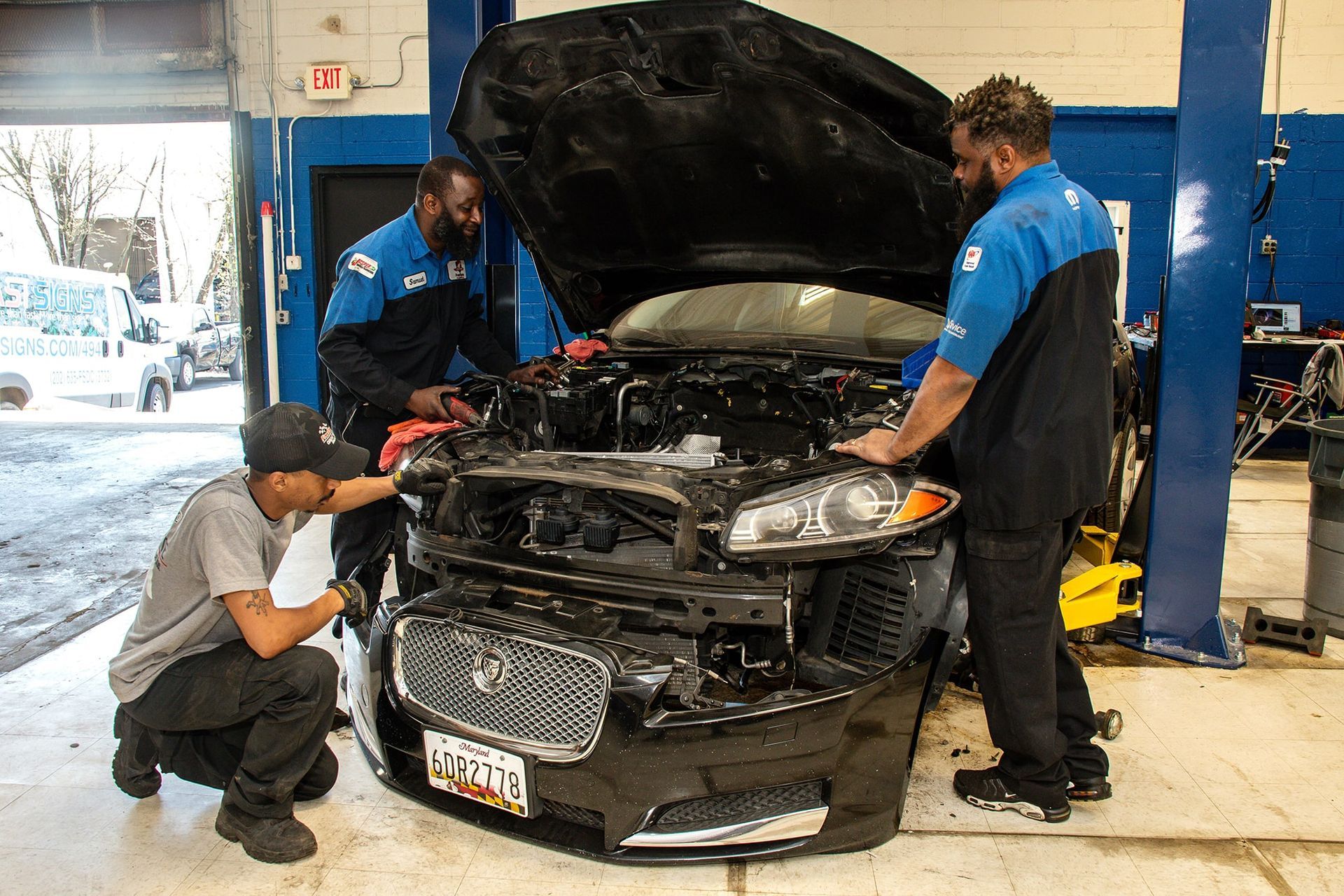 Three mechanics working on the front of a black Jaguar in a garage.