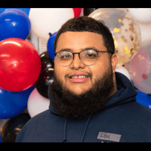 Man with a beard and glasses smiling in front of blue, red, and white balloons. He wears a blue hoodie.