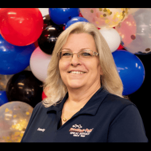Woman in glasses, polo shirt, smiling, in front of red, white, blue, black balloons.