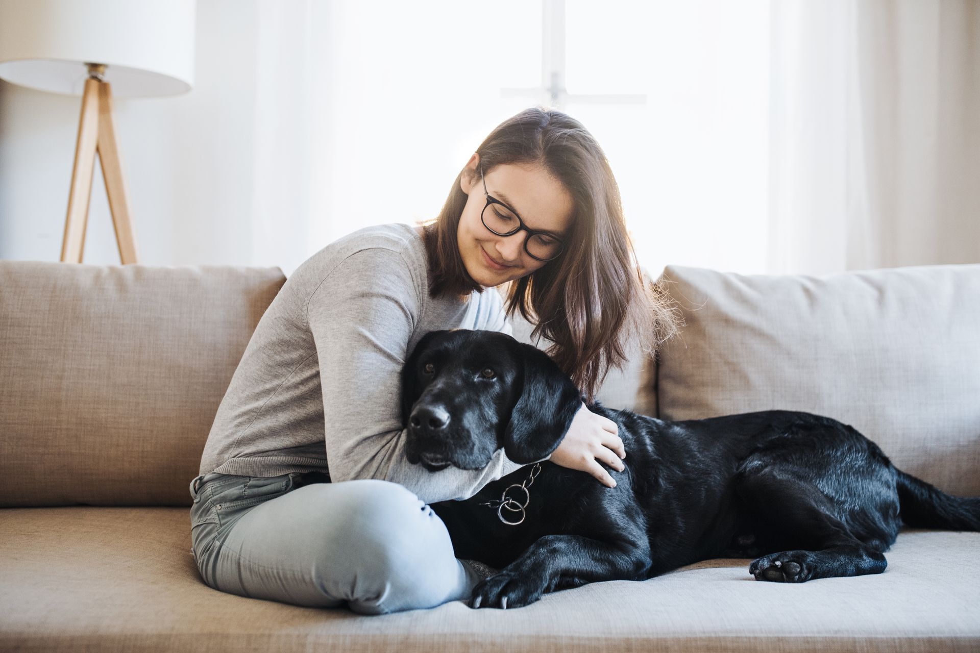 Woman wearing glasses pets a black dog on a beige couch in a sunlit living room.