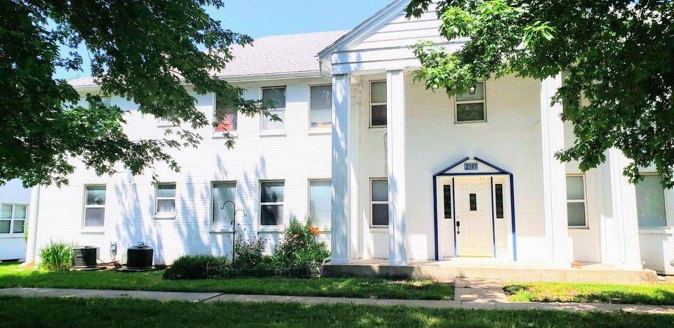 White building with columns, several windows, and a doorway, partially framed by green trees.