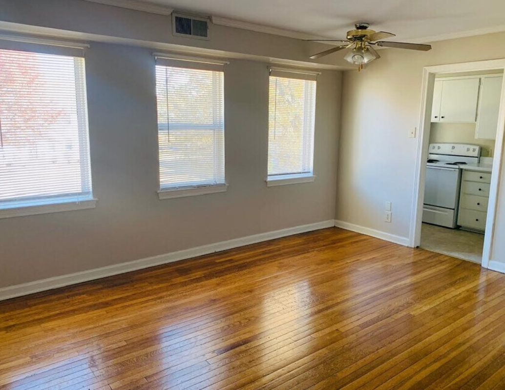 Empty room with hardwood floors, three windows with blinds, and a view into a small kitchen.