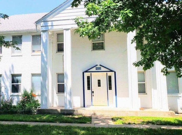White two-story building with large white columns flanking the front door; blue trim. Green foliage on the right.