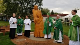 Priests in green vestments stand beside a large orange sculpture outdoors at a church ceremony