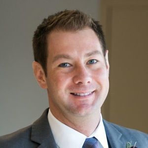 Smiling man in a gray suit and blue tie against a neutral indoor background