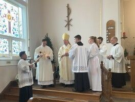Clergy in white robes stand at an altar during a church service near stained-glass windows.