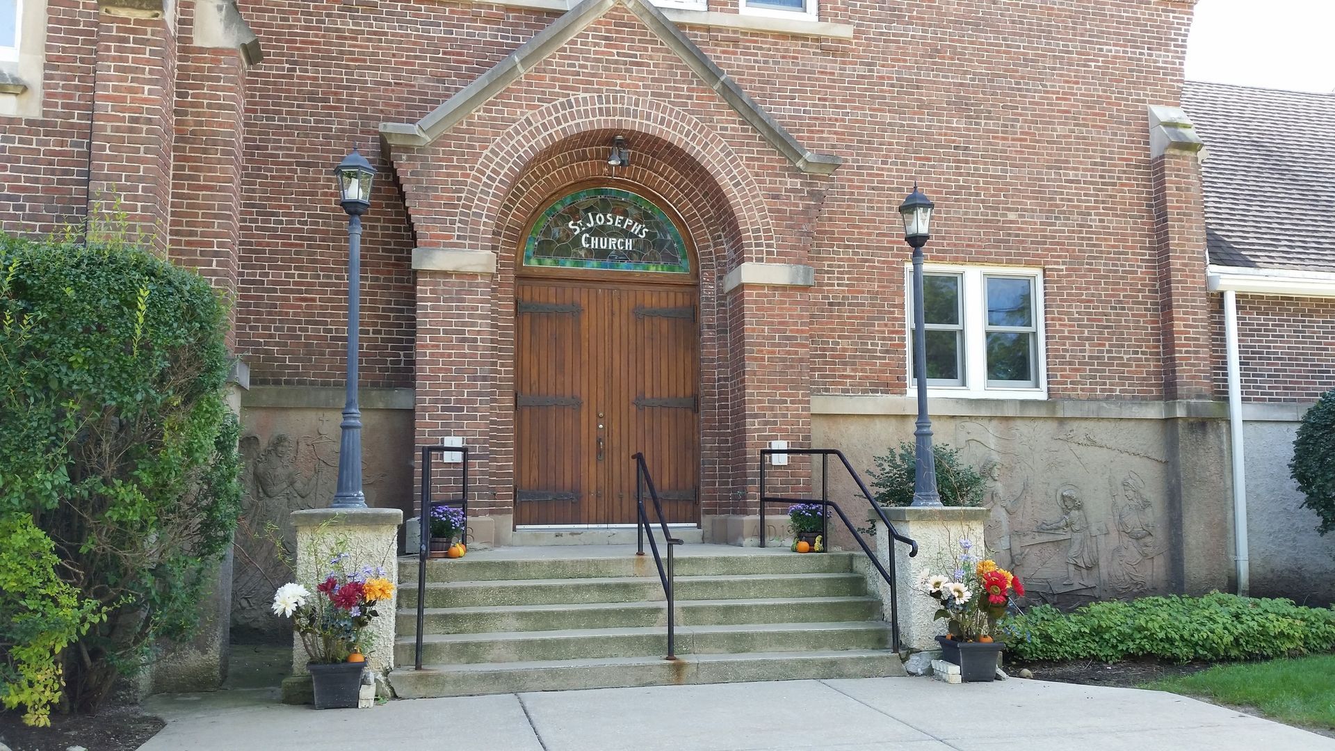 Brick building entrance with arched wooden door, steps, black railings, and potted flowers by the walkway