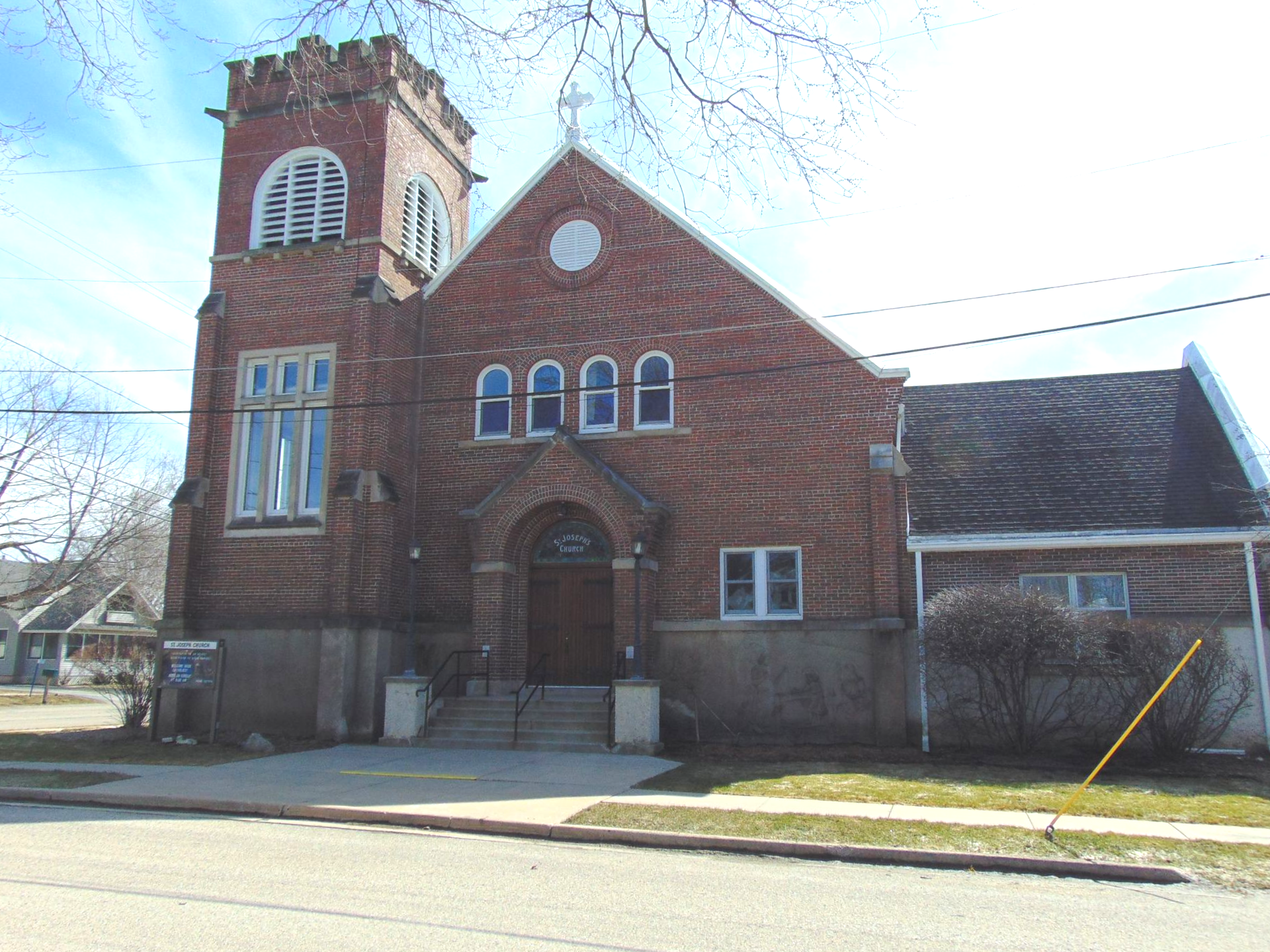Red brick church with a small tower and arched windows beside a sidewalk on a sunny day