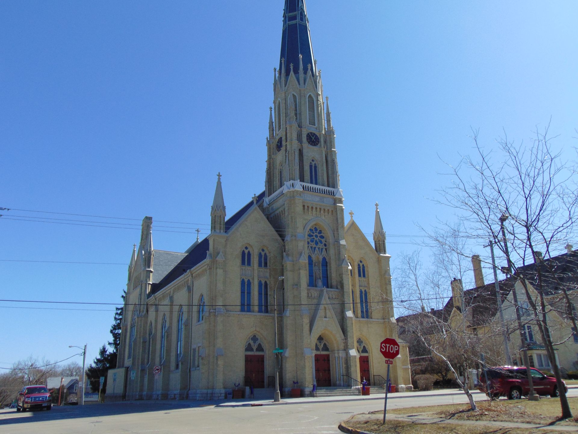 Stone church with a tall spire on a sunny street corner, viewed from the road.