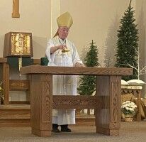 Priest in white and gold robes stands at church altar with trees in the background