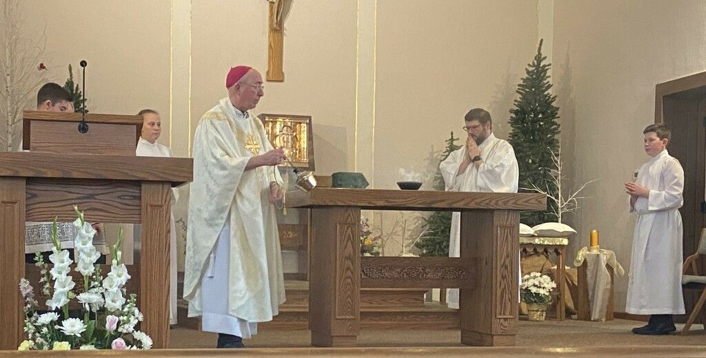 Clergy in white robes stand at the altar during a church service, with flowers and candles nearby